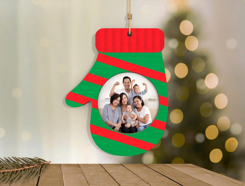 May include: A Christmas ornament shaped like a mitten, with a family photo in the center. The mitten is green with red stripes and a red top. A string is attached for hanging. The background is blurred with a Christmas tree.