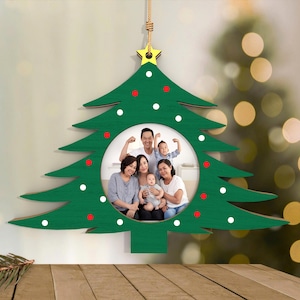 May include: A green wooden Christmas tree ornament with a circular photo frame. The tree is decorated with white and red dots and a gold star at the top. A family portrait is visible inside the frame. The ornament hangs from a natural-colored string.