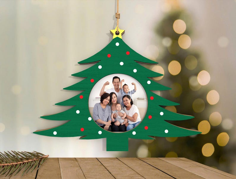 May include: A green wooden Christmas tree ornament with a circular photo frame. The tree is decorated with white and red dots and a gold star at the top. A family portrait is visible inside the frame. The ornament hangs from a natural-colored string.