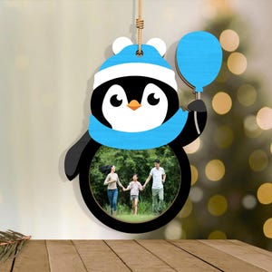 May include: A wooden penguin-shaped photo frame with a family photo inside. The penguin wears a blue hat and scarf and holds a blue balloon. The frame hangs from a natural-colored string, perfect for holiday decor.
