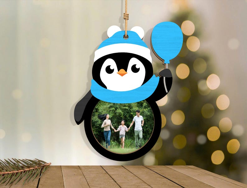 May include: A wooden penguin-shaped photo frame with a family photo inside. The penguin wears a blue hat and scarf and holds a blue balloon. The frame hangs from a natural-colored string, perfect for holiday decor.
