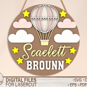 May include: A brown wooden circle with a hot air balloon design. The balloon is pink and white with yellow stars around it. The text "Scaelett Brounn" is written in white on the circle.