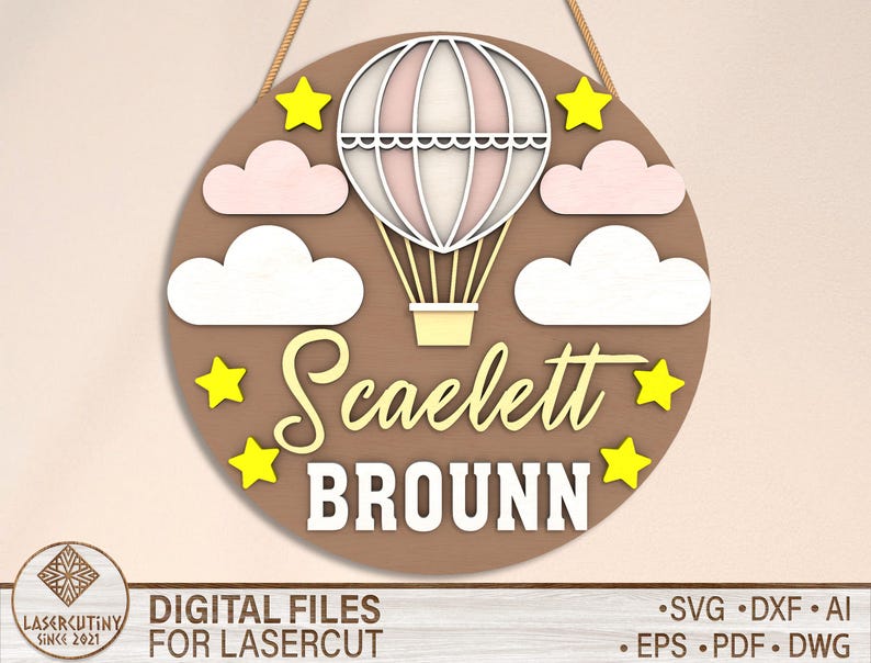 May include: A brown wooden circle with a hot air balloon design. The balloon is pink and white with yellow stars around it. The text "Scaelett Brounn" is written in white on the circle.