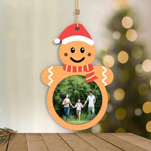 May include: Wooden gingerbread man Christmas ornament with a photo frame. The ornament features a red Santa hat, a red and white striped scarf, and a smiling face. The photo frame holds a family photo. The ornament hangs from a natural twine.