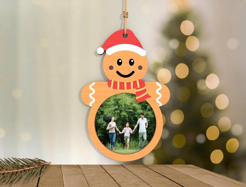 May include: Wooden gingerbread man Christmas ornament with a photo frame. The ornament features a red Santa hat, a red and white striped scarf, and a smiling face. The photo frame holds a family photo. The ornament hangs from a natural twine.