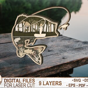 May include: A laser cut wooden design of a bass fish with a fisherman in a boat inside the fish. The design is layered and has a rustic wood finish. The text "DIGITAL FILES FOR LASER CUT 9 LAYERS" is at the bottom of the image.