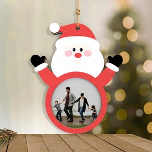 May include: A wooden Santa Claus ornament with a red hat and suit, white beard, and black gloves. The ornament has a circular opening for a photo. A family is pictured inside the frame, and the ornament hangs from a jute string.
