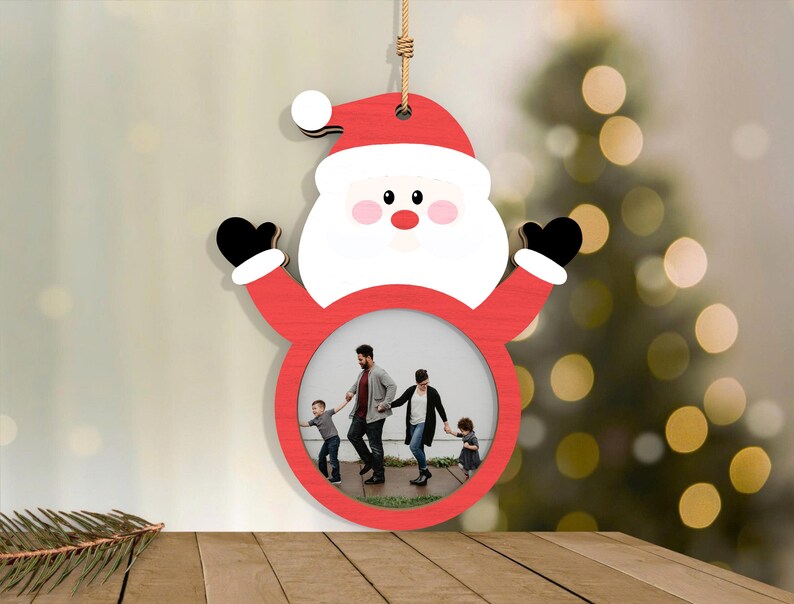 May include: A wooden Santa Claus ornament with a red hat and suit, white beard, and black gloves. The ornament has a circular opening for a photo. A family is pictured inside the frame, and the ornament hangs from a jute string.