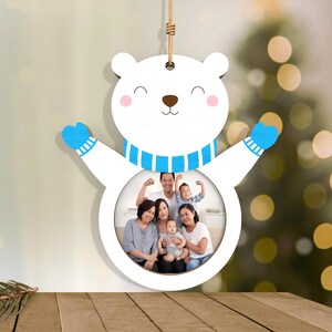May include: A white wooden polar bear ornament with a circular photo frame. The bear has blue mittens and a blue and white striped scarf. The photo frame contains a family portrait. A brown string is attached for hanging.