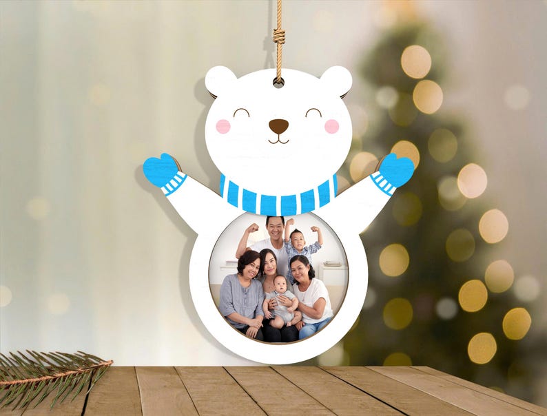 May include: A white wooden polar bear ornament with a circular photo frame. The bear has blue mittens and a blue and white striped scarf. The photo frame contains a family portrait. A brown string is attached for hanging.