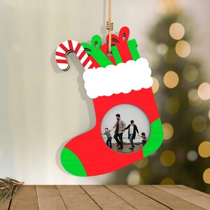 May include: A red and green Christmas stocking-shaped ornament with a white cuff and a candy cane. The ornament has a circular opening to display a photo. The stocking is hanging by a natural fiber rope. The background is blurred with a Christmas tree.