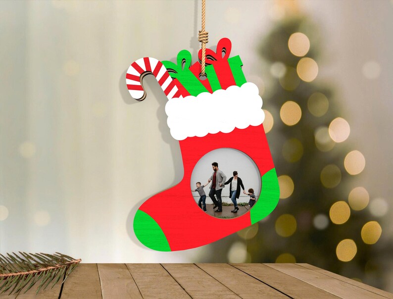 May include: A red and green Christmas stocking-shaped ornament with a white cuff and a candy cane. The ornament has a circular opening to display a photo. The stocking is hanging by a natural fiber rope. The background is blurred with a Christmas tree.