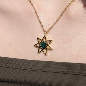 May include: Gold-toned necklace with a starburst pendant featuring a dark green center stone. The necklace is worn over a dark green tank top. The image is well-lit, highlighting the jewelry's details. The brand name "Emissary of Wind" is visible.