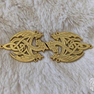 Puede incluir: Dos cierres metálicos dorados con un diseño vikingo. Cada cierre presenta un patrón simétrico de líneas entrelazadas y figuras de dragones. Los cierres están conectados por pequeños bucles, sobre un fondo blanco y esponjoso.