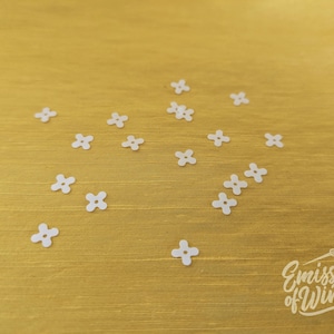 Peut inclure: Petits ornements blancs en forme de fleurs à quatre pétales dispersés sur une surface dorée. Les embellissements sont petits et espacés uniformément. Le fond est une surface dorée texturée. Le logo "Emissary of Wind" est en bas à droite.