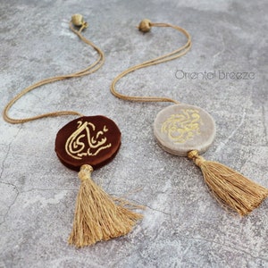 Może przedstawiać: Dwa okrągłe ozdoby z aksamitu z haftem złotym i frędzlami. Ozdoby są brązowe i szare i wiszą na sznurku.