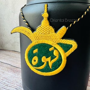 May include: A gold and green embroidered patch with Arabic script in a circle, attached to a black container with a gold and black cord. The text on the patch reads "Oriental Breeze".
