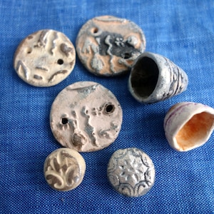 Può includere: Una collezione di perline e ciondoli in ceramica fatti a mano in varie forme e dimensioni. I pezzi sono principalmente bianco sporco con tocchi di grigio, marrone e rosa, e presentano disegni e motivi strutturati. Gli articoli sono esposti su una superficie di tessuto blu.