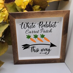 以下が含まれることがあります： 黒い筆記体で「White Rabbit Carrot Patch this way」と書かれた木製の看板。看板は茶色の木製の枠で、緑の葉が付いたオレンジ色のニンジンが3本描かれている。