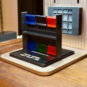 May include: A black hat clip jig with a rectangular base and a vertical frame holding six colored clips: blue, black, red, blue, green, and red. The text "FLIP3 30MM HAT CLIP JIG" is printed on the base.