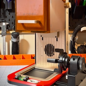May include: An xTool laser machine featuring a translucent amber protective cover, a vertical silver metal frame with a central fan, and a red base. The unit includes a black rotary attachment on the right side and a small green spirit level.
