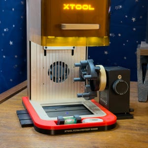 May include: A laser engraving machine with the text "XTOOL" on top. The machine has a red and silver base, a gold-colored top, and a black rotary attachment. The text "XTOOL F2 RA3 ROTARY RISER" is visible on the base.