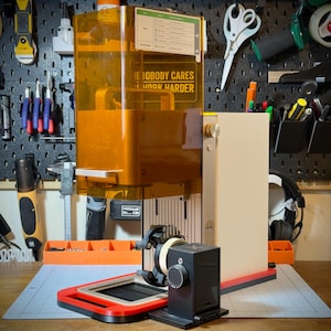 May include: A resin 3D printer featuring a tall, translucent amber-colored protective cover, a cream-colored vertical body, and a black base with a red trim. A small black control module with a circular dial is attached to the front of the machine.