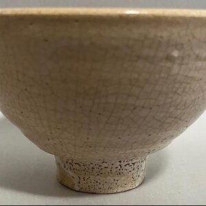 Könnte beinhalten: Eine beige Keramikschale mit einer breiten, abgerundeten Form und einem kleinen, strukturierten Fuß. Die Oberfläche der Schale weist ein Craquelé-Muster auf, das ihr ein einzigartiges, gealtertes Aussehen verleiht. Die Schale wird wahrscheinlich zum Servieren von Speisen verwendet.