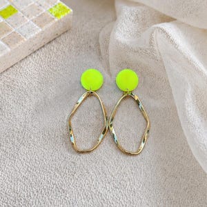 May include: A pair of gold-toned, irregular-shaped dangle earrings with neon green circular studs. The earrings are displayed on a textured, light-colored surface with a white fabric backdrop.