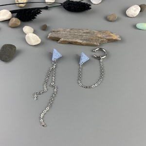 Könnte beinhalten: Asymmetrische Ohrringe mit hellblauen Dreieckssteckern und silbernen Ketten. Ein Ohrring ist ein Stecker mit einer hängenden Kette, der andere ist ein Ohrklemme mit einer Kette. Die Ohrringe liegen auf einer grauen Oberfläche mit dekorativen Steinen und getrockneten Pflanzen.