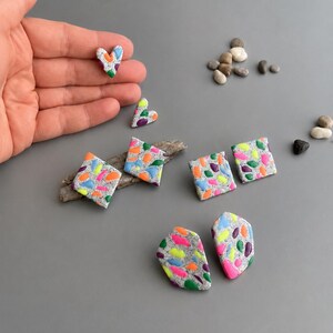 Op de afbeelding: Handgemaakte polymeerklei oorbellen en hangers in hart- en vierkantvormen. De grijze stukken zijn bespikkeld met neonroze, geel en blauw. Sommige stukken zijn op drijfhout geplaatst.