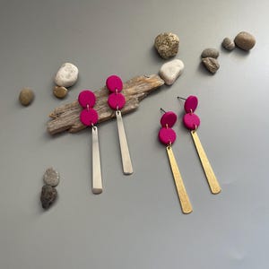 Könnte beinhalten: Ein Paar Ohrringe mit zwei rosa Tonkreisen und einem langen silbernen Anhänger, und ein weiteres Paar mit zwei rosa Tonkreisen und einem langen goldenen Anhänger.