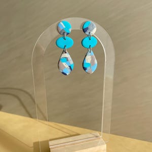 Könnte beinhalten: Ein Paar Ohrhänger, präsentiert auf einem transparenten Acrylständer. Jeder Ohrring hat eine kreisförmige Oberseite mit einem blauen, grauen und weißen Muster, ein türkisfarbenes Oval und eine Tropfenform mit einem ähnlichen Muster.