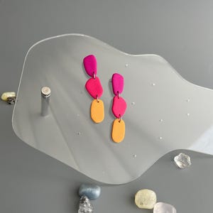 Könnte beinhalten: Ein Paar abstrakte Hängeohrringe. Jeder Ohrring hat drei farbige, unregelmäßige Formen in Fuchsia, Pink und Orange. Die Ohrringe werden auf einem klaren, wellenförmigen Ständer vor grauem Hintergrund präsentiert.
