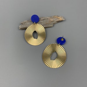 Könnte beinhalten: Ein Paar goldfarbene, strukturierte Ohrringe mit offenem, kreisförmigem Design. Jeder Ohrring hat einen kleinen, blauen runden Stud an der Oberseite. Die Ohrringe werden auf einer grauen Oberfläche präsentiert.