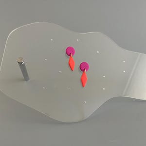 Könnte beinhalten: Ein Paar handgefertigte Ohrringe. Jeder Ohrring hat einen kleinen, runden, magentafarbenen Stecker, der mit einem leuchtend orangefarbenen, rautenförmigen Anhänger verbunden ist. Die Ohrringe werden auf einem klaren, gebogenen Präsentationsständer gezeigt.