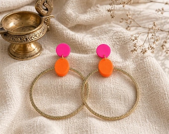 Puede incluir: Un par de pendientes de aro dorados con un diseño texturizado, cada uno con un círculo rosa brillante sobre un círculo naranja vibrante. Los pendientes se exhiben sobre una tela de color crema, con un cuenco de latón decorativo y flores secas en el fondo.