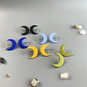 Könnte beinhalten: Ohrstecker in Form von Halbmonden in Schwarz, Blau, Gelb und Grün. Die Ohrringe werden auf transparenten Kunststoffständern präsentiert, mit kleinen Edelsteinen daneben. Ein einzigartiges Accessoire, das jedem Outfit einen Hauch von Mondzauber verleiht.