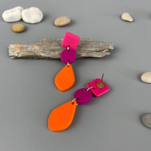 Op de afbeelding: Een paar oorhangers met geometrische vormen. De oorhangers zijn gemaakt van polymer clay en hebben een roze vierkant, een roze cirkel, een oranje traan en een kleinere oranje traan. De oorhangers zijn bevestigd aan zilveren oorhaken.