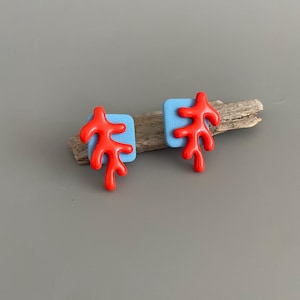 Könnte beinhalten: Ohrstecker mit einem leuchtend roten Korallenmuster auf einem hellblauen Quadrat. Die Ohrringe werden auf Treibholz vor grauem Hintergrund präsentiert. Ein einzigartiges Accessoire, das jedem Outfit einen Farbtupfer verleiht.