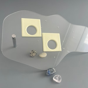 Könnte beinhalten: Ein klarer Acryl-Displayständer mit zwei quadratischen, cremefarbenen Einsätzen, jeweils mit einem kreisförmigen Ausschnitt. Ein kleiner, silberner Metallstab und mehrere kleine Steine in verschiedenen Farben sind ebenfalls vorhanden.