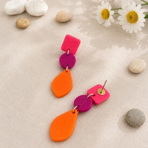 Peut inclure: Une paire de boucles d'oreilles faites à la main. Chaque boucle d'oreille présente un carré, un cercle et une forme de goutte dans des tons de rose, de violet et d'orange. Les boucles d'oreilles sont posées sur un fond neutre avec de petites pierres et des marguerites.