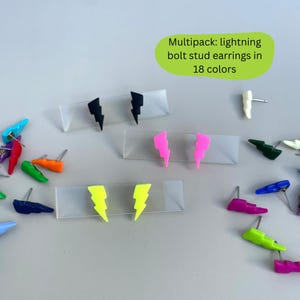 Op de afbeelding: Een verzameling kleurrijke bliksemschicht oorknopjes in verschillende kleuren, waaronder zwart, roze en geel. De afbeelding bevat de tekst "Multipack: bliksemschicht oorknopjes in 18 kleuren."