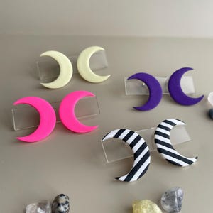Könnte beinhalten: Eine Auswahl an halbmondförmigen Ohrringen in verschiedenen Farben und Mustern. Die Ohrringe umfassen Volltonfarben wie Gelb, Pink und Lila sowie ein schwarz-weiß gestreiftes Design. Kleine, transparente Präsentationsständer halten die Ohrringe.