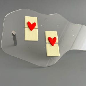 Könnte beinhalten: Transparente Acryl-Ausstellung mit zwei cremefarbenen rechteckigen Elementen, die jeweils mit einem roten Herzen versehen sind. Eine silberne Metallstange ist an der Ausstellung befestigt. Der Hintergrund ist einfarbig grau.