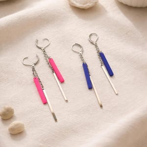 Könnte beinhalten: Silberfarbene Ohrhänger mit einem Stangendesign. Ein Paar hat einen leuchtend pinkfarbenen Stab, das andere einen blauen Stab. Die Ohrringe hängen an einer Kette und einem kleinen Haken.