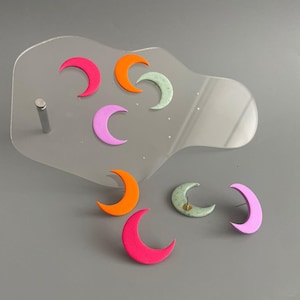 Könnte beinhalten: Ohrringe in Form von Halbmonden in verschiedenen Farben wie Pink, Orange und Grün, präsentiert auf einem transparenten Acrylständer. Die Ohrringe sind auf einem grauen Hintergrund angeordnet, der ihre lebendigen Farben hervorhebt.