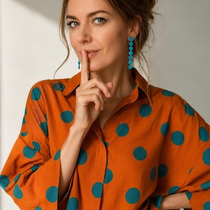 Puede incluir: Una camisa de manga larga con botones en color naranja quemado que presenta un patrón repetitivo de grandes lunares de color verde azulado. La camisa se combina con jeans azules y aretes largos y colgantes que consisten en una fila vertical de pequeños círculos de color verde azulado.