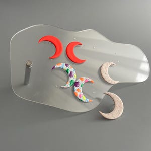 Könnte beinhalten: Ein klarer Acrylständer hält sechs halbmondförmige Objekte. Zwei sind leuchtend rot, zwei sind mit bunten Punkten gemustert und zwei sind gesprenkelt beige. Der Ständer hat einen silbernen Metallpfosten.