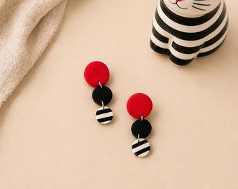 Peut inclure: Une paire de boucles d'oreilles pendantes à rayures rouges, noires et blanches. Les boucles d'oreilles comportent un cercle rouge en haut, suivi d'un cercle noir et d'un cercle blanc à rayures noires. Une figurine de chat rayé noir et blanc est en arrière-plan.